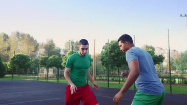Motivated sporty attractive Middle Eastern man basketball player dribbling ball against black male opponent, giving pass to teammate while multiethnic athletes playing streetball on outdoor court.