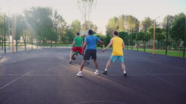 Skillful sporty male basketball player making assist to African American man teammate to score two points with layup shot, while multiracial athletes playing streetball game on outdoor court.
