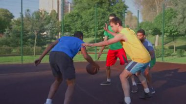 Motivated sporty fit black male basketball player receiving pass from teammate, making field goal attempt to score two points while multiracial athletes playing streetball game outdoors.