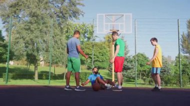 Sporty fit Middle Eastern male streetball player extending hand to lift fallen black opponent player off ground, helping to stand up, showing good sportsmanship during basketball game on outdoor court