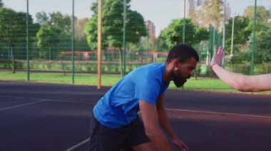Portrait of skillful active sporty fit African American male basketball player receiving pass from teammate, making three points shot while multi ethnic athletes playing streetball on outdoor court.
