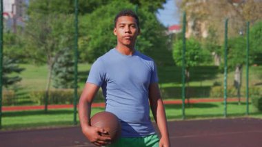 Ciddi bir Afro-Amerikan erkek basketbolcunun portresi, topu elinde tutuyor, açık sahada duruyor, kararlılığını ve güvenini gösteriyor..