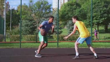 Sportif formda siyah basketbolcu potaya doğru gidiyor, tek elle smaç vurduktan sonra sayı yapıyor, dışarıda sahanın üzerinde sokak topu oynayan çok ırklı spor takımları..