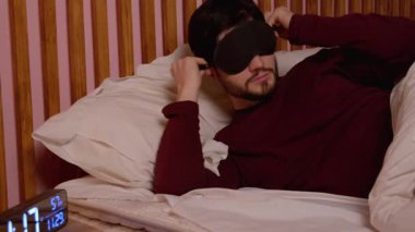 Çekici, sakallı, orta yaşlı, pijamalı, ipek uyku maskesi takan, geceleri rahat bir yatakta dinlenirken iyi bir uyku ve restorasyon için hazırlanan bir adam..