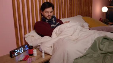Endişeli, yakışıklı, sakallı, olgun bir adam, sıcak bir eşarba sarılmış, yatağa oturmuş, ateş ve grip virüsünden muzdarip, ölçüm yapıyor, yatak odasında dijital termometreyle sıcaklığı ölçüyor..