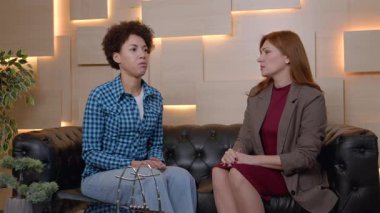 Sempatik çekici olgun kadın akıl sağlığı uzmanı koltukta oturuyor, depresif siyah kadın hastayı rahatlatıyor, psikoterapi seansı sırasında anlayış, ilgi ve destek gösteriyor..