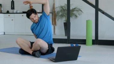 Fitness minderinde bacak bacak üstüne atmış, yoga pozisyonu almış, yoga dersini ev içi spor eğitimi sırasında laptopta takip eden motive olmuş sportif formda bir adam..