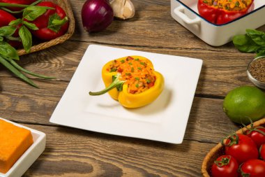 Et ve sebzeyle doldurulmuş dolmalık biberler, eski bir kırsal masada beyaz kare bir tabakta peynirle pişirilmiş. Kapat..