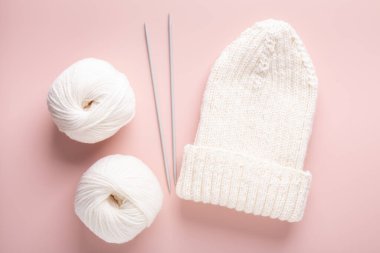 Beyaz yün örgü iğneleri ve pastel pembe arka planda örülmüş şapka. Kadın örgü hobisi. Üst görünüm