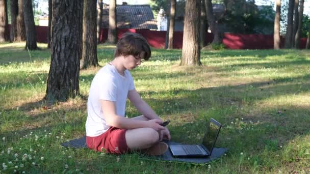 Unge man sitter på gräset i parken med sin laptop och pratar i telefon. Begreppet distansutbildning — Stockvideo