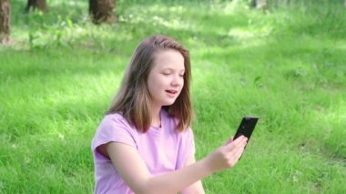 Akıllı telefonu olan genç bir kız video görüşmesi yapıyor. Şehir parkında çimlerde oturan ve cep telefonuyla arkadaşlarıyla konuşan bir kız.