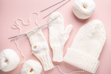 Beyaz yün demeti, örgü iğneleri, şapka ve eldivenler pastel pembe arka planda örgü işleme sürecinde. Kadın hobisi. Üst görünüm