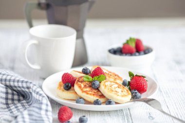 Taze dutlu süzme peynir krepleri, beyaz ahşap masada bir fincan kahve makinesi. Lezzetli kahvaltı yemekleri. Syrniki.