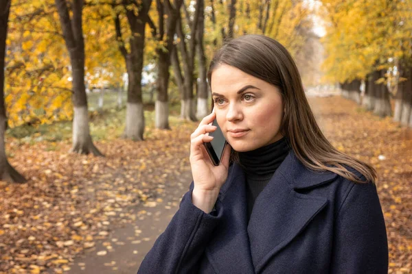 stock image Young woman in an autumn park with a smartphone. Girl talking on her mobile cell phone. Technology, communication and lifestyle concept