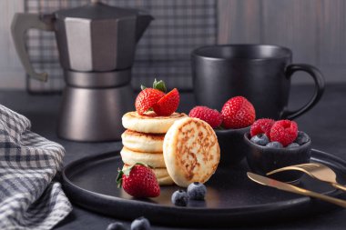 Koyu seramik tabakta taze çilek ve yabanmersinli süzme peynirli krep. Bir fincan kahve ve kahve makinesi.