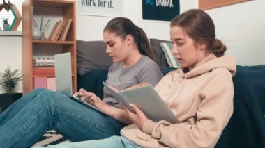 Genç kolej öğrencilerinin yerde oturup ödev yaparken orta boy fotoğrafları. Bir tanesi, arkadaşı bilgisayarda yazarken ders kitabından yüksek sesle okuyor.