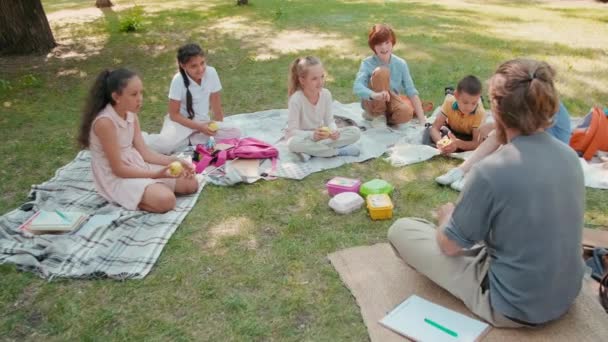 跟踪坐在公园毯子上的放大镜的男教师慢镜头 并与一群听他讲话的学生交谈 课后吃午饭 — 图库视频影像