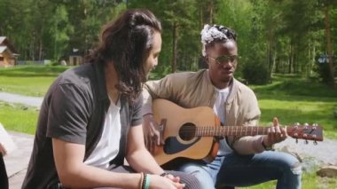 İki etnik çeşitlilikte çocuğun yazın dışarıda oturup şarkı söyleyip gitar çalarak atmosferin tadını çıkardığı ve dinlendiği orta büyüklükte görüntüler.