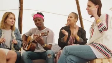 Gün ışığında kamp alanında beyaz çadırda oturup gitar melodisi çalan neşeli genç yetişkinlerin orta ölçekli görüntüsü.