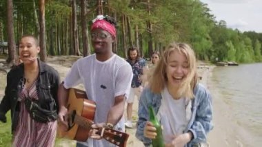 Yakışıklı Afrikalı Amerikalı müzisyen ve arka planda konuşan çiftin kahkahalarla gitar çalması arasında geçen genç ve güzel kadınların yavaş çekim görüntüleri.