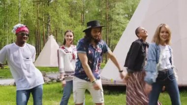 Yavaş çekim, genç neşeli arkadaş grubunun el ve bacaklarıyla dans ederek kamp alanında sırt çantalı çocuk dansı yapmasını gösteren orta ölçekli görüntüler.