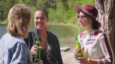 Gün ışığında göl kıyısında dikilmiş içki şişeleriyle konuşan ve gülen genç kadınların orta boy görüntüleri.