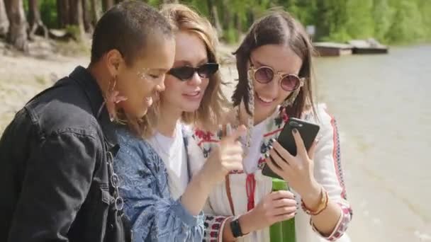 携帯電話の画面を見ている流行の若い女性の中規模の閉鎖感情的に日光の湖畔に立って写真を議論 — ストック動画