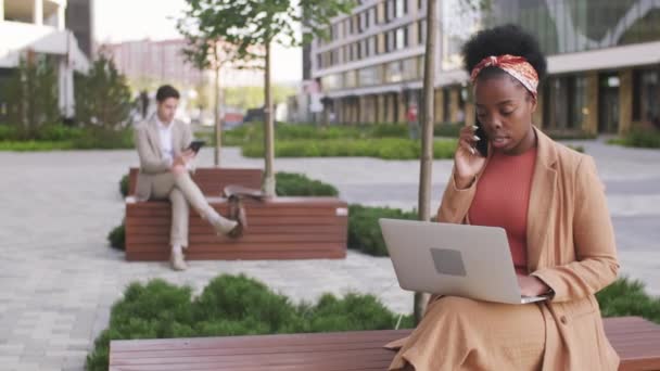 幸せなアフリカ系アメリカ人ビジネスマンのショットのシーケンスビジネスセンターの外のベンチに座って ノートパソコンで作業しながら携帯電話で話す — ストック動画