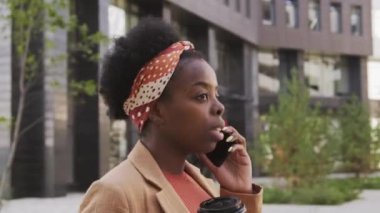  Meşgul siyahi iş kadınının, kahve bardağında kahve taşıdığı ve dışarıda yürürken cep telefonuyla konuştuğu yan görüntüler.