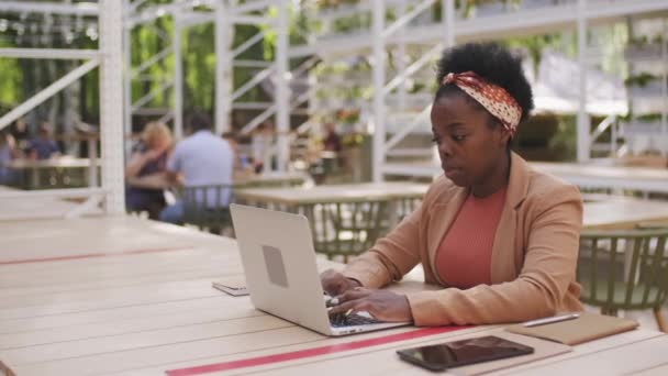カフェの屋外パティオにテーブルに座ってノートパソコンで作業忙しい黒人実業家の追跡ショット — ストック動画
