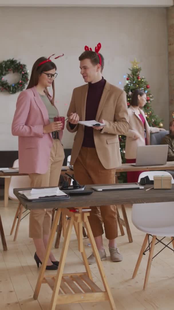 圣诞当天 年轻快乐的女商人和戴着节日帽带的商人在办公室聊天时的垂直镜头 — 图库视频影像