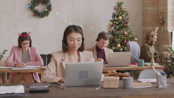 PAN ázsiai üzletasszony fülhallgatóban ül az asztalnál nyitott terű irodában és csevegik valakivel, miközben dolgozik karácsonykor. Díszített karácsonyfa és villogó tündér fények a háttérben