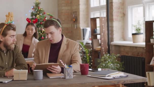 スタートアップ企業のオフィスのデスクに座ってクリップボード上の文書を議論しながら クリスマスに取り組んで忙しい若いビジネスマンのPan — ストック動画