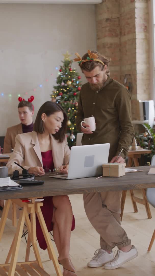 一个留着胡子的商人头戴喜庆的头巾 站在他的女同事旁边 一边看着笔记本电脑屏幕一边讨论圣诞节的工作 一边垂直地拍照 — 图库视频影像
