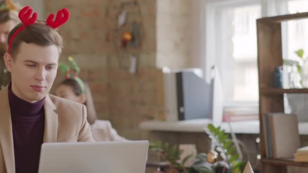 Noel Yoğun Bir Ofiste Dizüstü Bilgisayarda Çalışan Neşeli Boynuzlu Genç — Stok video