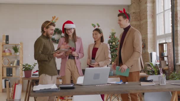 クリスマスの日にオフィスでお祝いのヘッドバンドやサンタの帽子チャットで幸せなスタートアップ企業の従業員の中ショット 背景に飾られたクリスマスツリー — ストック動画