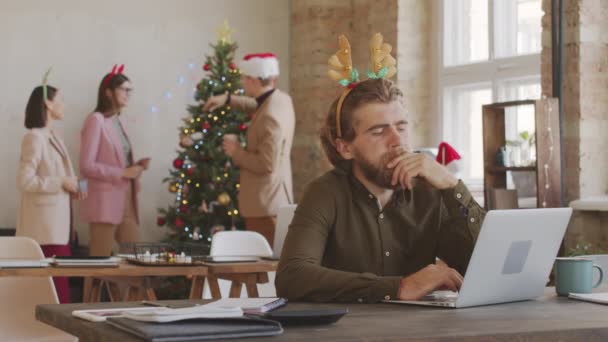 クリスマスのオフィスでラップトップで働いているお祝いのヘッドバンドでストレスのある男の中のショット 彼は疲労で目をこすり クリスマスツリーでチャット彼の幸せな同僚として画面を見ている — ストック動画