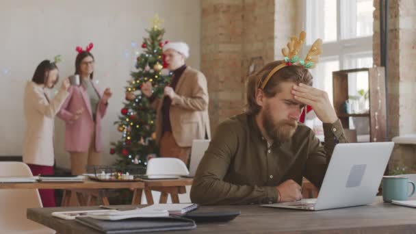 圣诞期间 一位留着喜庆头巾的大胡子商人坐在办公桌前 一边看笔记本电脑 一边拍了一张中等比例的照片 快乐的同事在圣诞树旁聊天跳舞 — 图库视频影像
