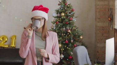 Gözlüklü mutlu genç iş kadınının portresi ve Noel Baba şapkası Noel için süslenmiş olarak ofiste duruyor ve fincandan kahve içiyor, sonra kameraya bakıp gülümsüyor.