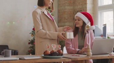 Asyalı bir kadın Noel Baba şapkasıyla bayan meslektaşına doğru yürüyor ve ona Noel hediyesi veriyor. Birbirlerine sarılıp tebrik ediyorlar. Arka planda peri ışıkları olan Noel ağacı