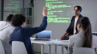 IT hocasının modern sınıfta programların kodlanması ve arka planda ekran projektörü ile ilgili ders materyallerini açıklarken orta boy görüntüsü ve öğrencilerden biri soru sormak için elini kaldırıyor.