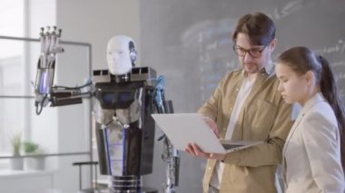 Genç modern öğretmenin, kadın ırk öğrencisinin, insansı robotlara uzaktan kumanda olarak dizüstü bilgisayarı kullanarak komutları nasıl takip ettirdiğini anlattığı orta büyüklükte görüntüler.