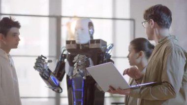 Genç erkek robotik profesörünün, insan benzeri elektronik robotlar için komut yazma işlemleri sırasında öğrencilerin önünde dizüstü bilgisayarda hareketler yapması.