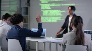 IT dersi sırasında bilgisayar sınıfında erkek öğrenci el kaldırıyor. Öğretmenlere bilgisayar programlama ve program kodlaması hakkında sorular soruyor.