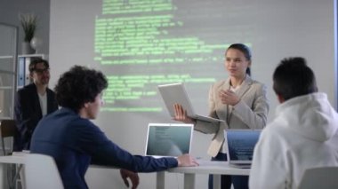Genç karışık ırk bilişim öğrencisi bilgisayar programında sunum yaparken, profesör dinlerken ve sınıf arkadaşları dizüstü bilgisayarlarla ekran projektörüne bakarken...