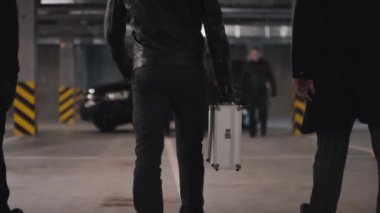 Gangster liderinin metal evrak çantasıyla diğer çok ırklı gangsterlere yürüyüp yeraltı otoparkında anlaşma yapmaya hazırlandığı görüntüler.