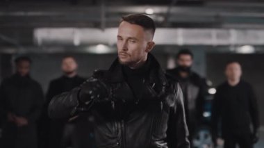 Deri eldivenli ve ceketli vahşi beyaz sakallı gangsterin portresi. Siyah güneş gözlüklerini çıkarıyor. Arka planda çetenin olduğu kameraya ciddi ciddi bakıyor.