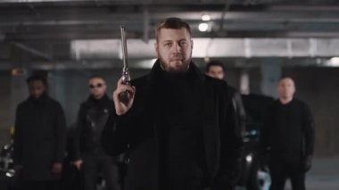 Orta boy, sakallı, beyaz, tabancalı, kameralı, sakız çiğneyen, siyah ceketli ve güneş gözlüklü bir haydudun yeraltı otoparkının arkasında durduğu görüntüler.