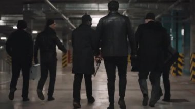 Arka planda vahşi, iddialı haydutların yeraltı otoparkında yürürken, tartışırken ve etraflarına gergin bir şekilde bakınırken görüntüler var.