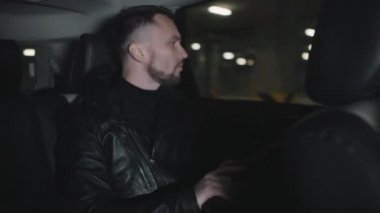 Arabasının arka koltuğunda oturan, kar maskesi takan ve silahlarını doldurup pencereden dışarı bakan vahşi, yakışıklı beyaz suçlunun orta boy yakın çekimi.